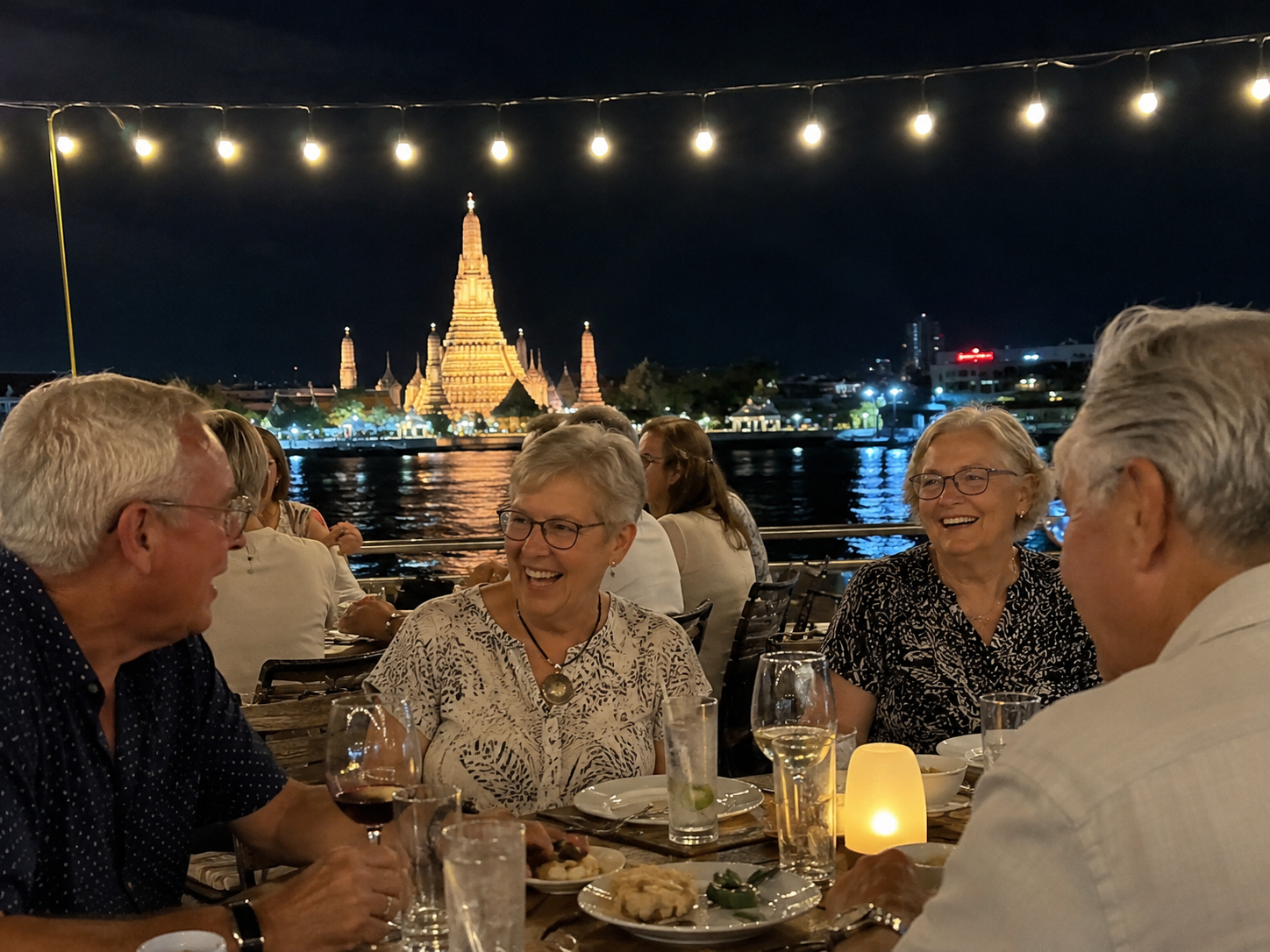New friends sharing dinner on a Chao Phraya river cruise in Bangkok, with Wat Arun behind them.